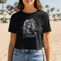 Black crop t-shirt with Chicana skull and rose graphic on a beach.