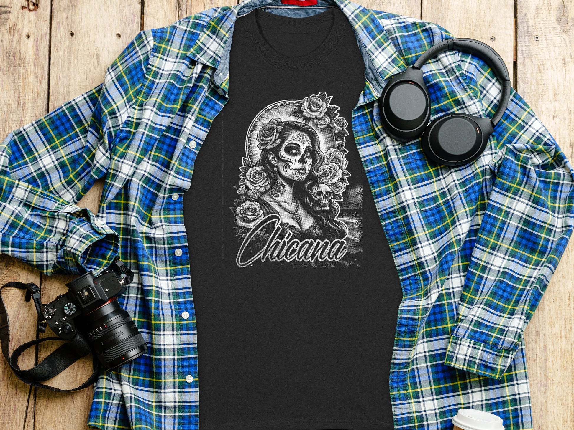 T-shirt with sugar skull artwork and "Chicana" text, laid flat with shirt, headphones, and camera.