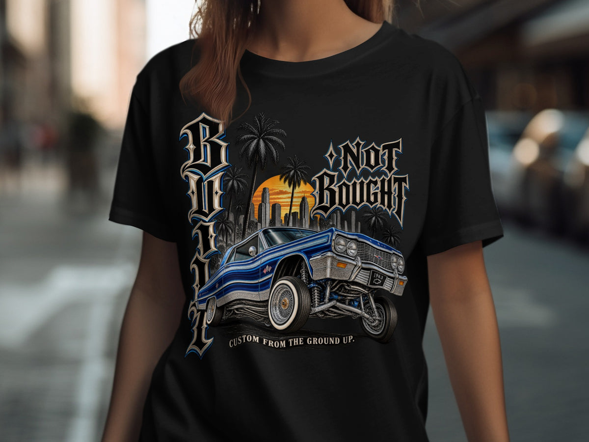 Black t-shirt with blue lowrider, city skyline, palm trees, and "NOT BOUGHT CUSTOM FROM THE GROUND UP." text.