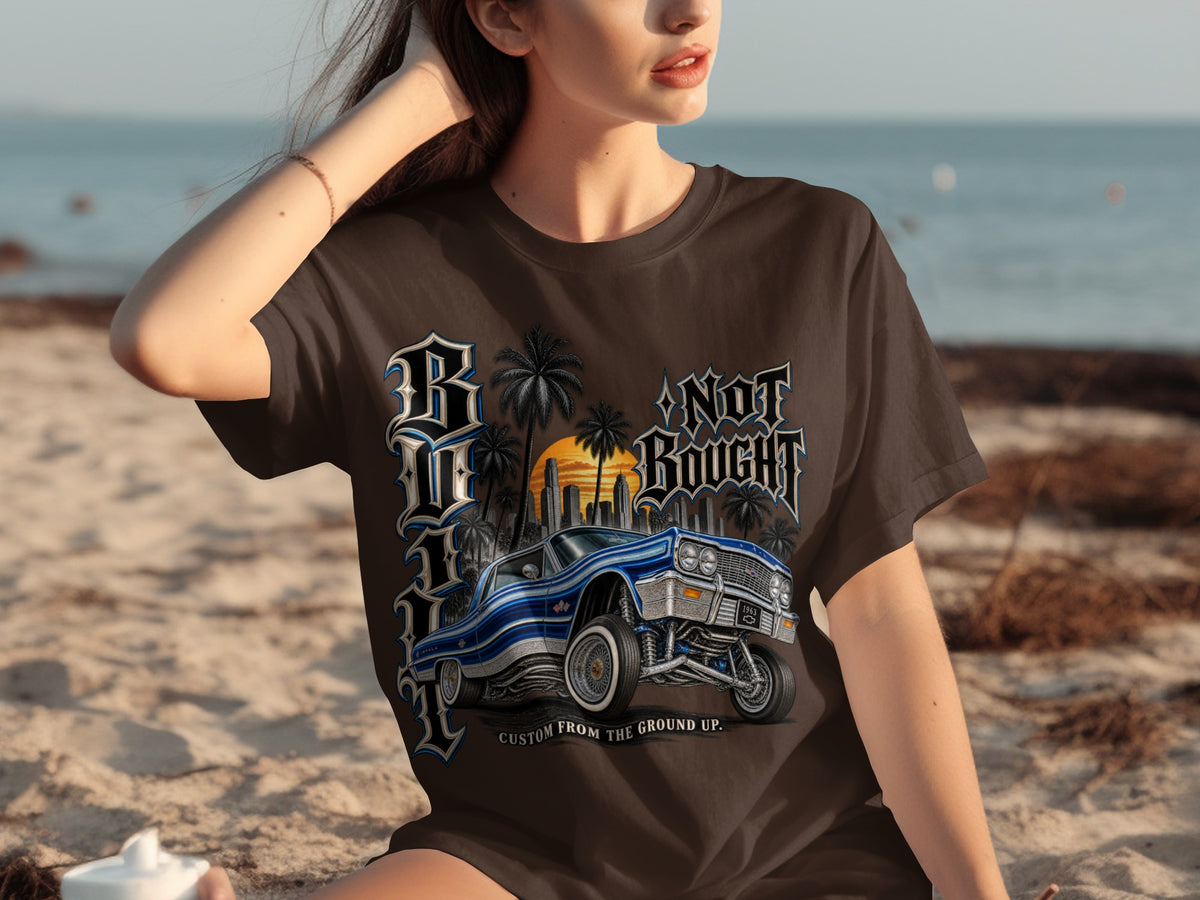Brown t-shirt with graphic of a blue classic car, palm trees, sunset, and text.