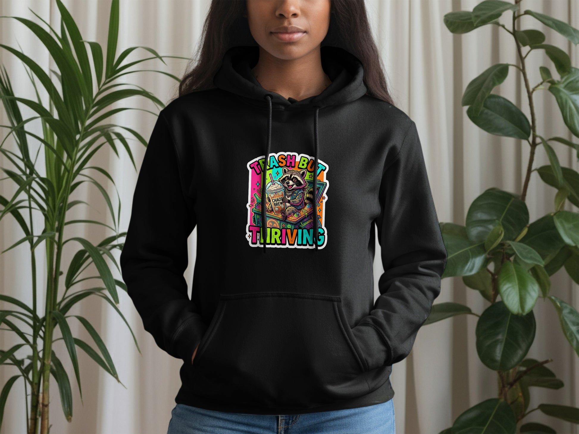 Black hoodie with "trash but thriving" graphic featuring raccoon, coffee, and plants.