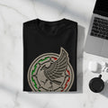 Black t-shirt with Mexico national football team crest, placed on a marble surface next to a laptop and earbuds.
