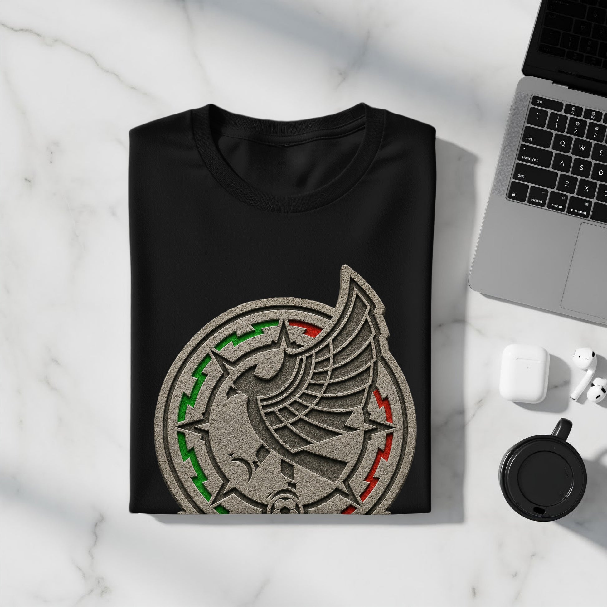 Black t-shirt with Mexico national football team crest, placed on a marble surface next to a laptop and earbuds.