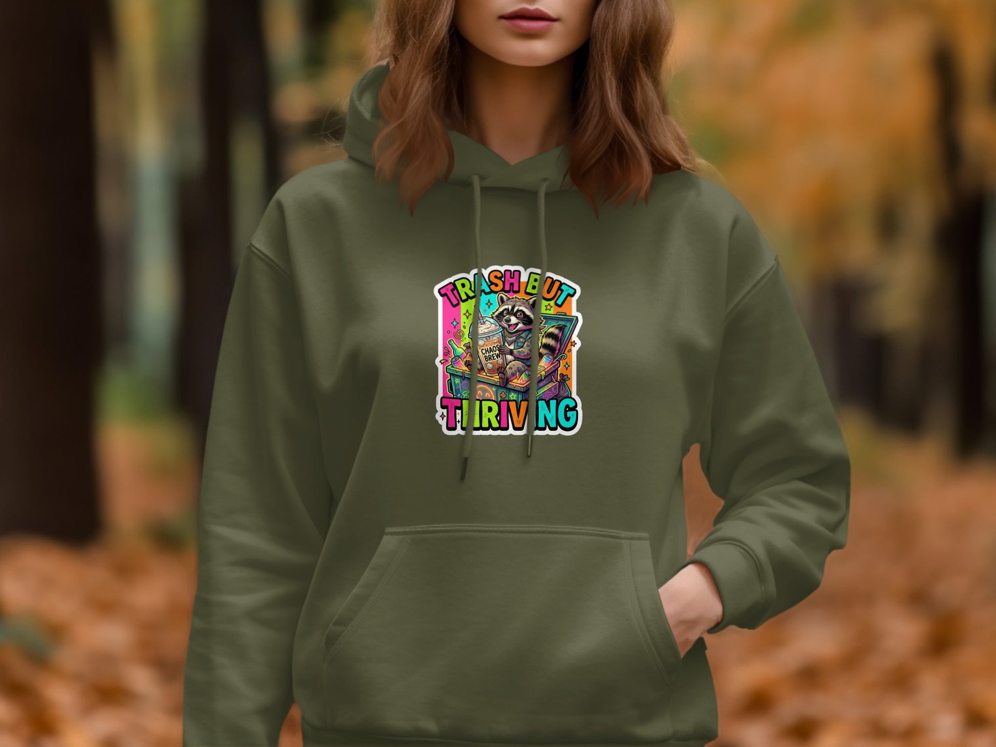 Olive green hoodie with raccoon, coffee, and "Trash but thriving" text graphic.
