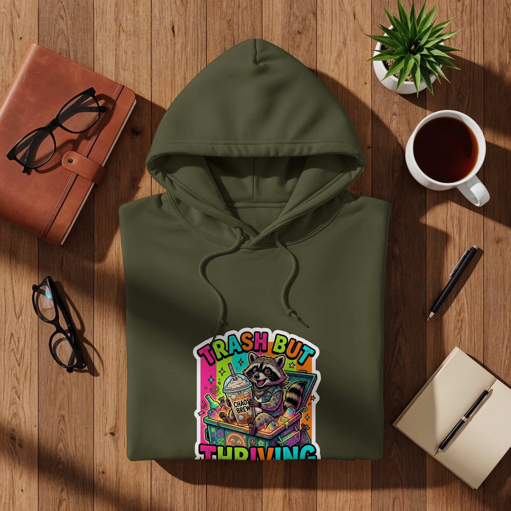 Green hoodie with "TRASH BUT THRIVING" raccoon graphic, on a wooden surface.