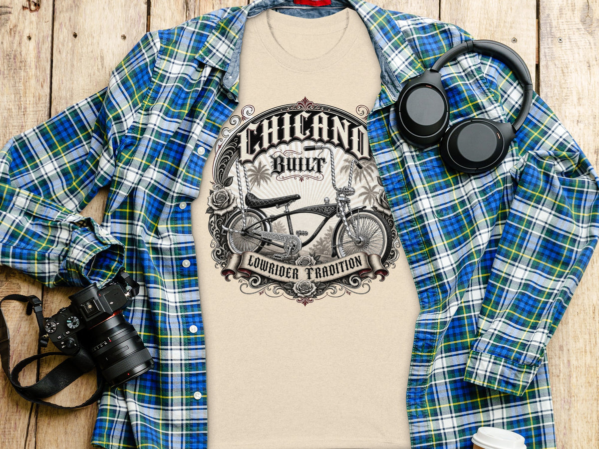 Tan t-shirt with "Chicano Built Lowrider Tradition" graphic, plaid shirt, headphones, camera